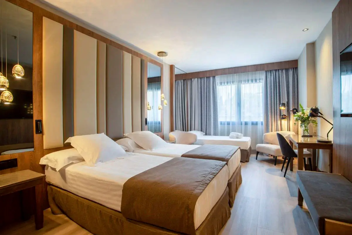 Habitación del hotel Granada Center donde se muestran dos camas unidas y una tercera cama supletoria al fondo junto a la ventana. También existe escritorio, silla y sofá.